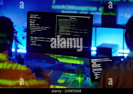 Computer screen with decoded data and two young contemporary programmers sitting by desks and developing new programs Stock Photo