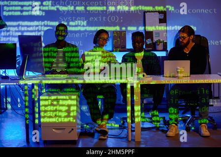 Row of four young intercultural diversity programmers sitting by desk in front of laptops during presentation of new software Stock Photo