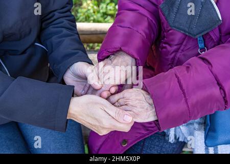 A mature woman holds her elderly mother's hands as they sit on a bench Stock Photo