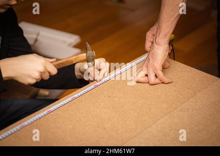 Close up of male hands measuring length of plywood with tape measure and female hands hammering nail into cheapboard. Assembling piece of furniture at Stock Photo