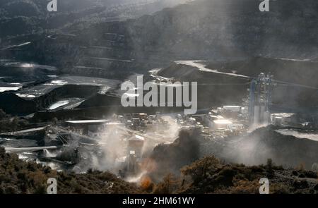 Mining plant in stone quarry in clouds of dust from production, backlit dramatic landscape with sun flare Stock Photo
