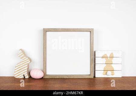 Mock up wood frame with Easter decor on a wood shelf. Shabby chic wood sign, egg, bunny. Square frame against a white wall. Copy space. Stock Photo