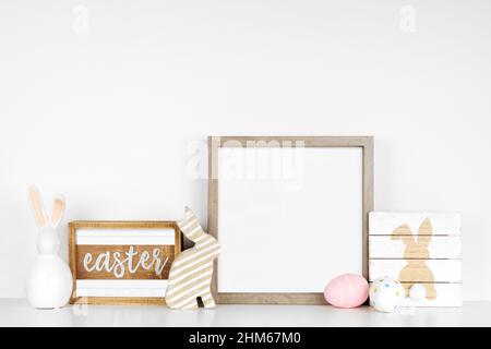 Mock up wood frame with Easter decor on a wood shelf. Shabby chic wood signs, eggs, bunnies. Square frame against a white wall. Copy space. Stock Photo