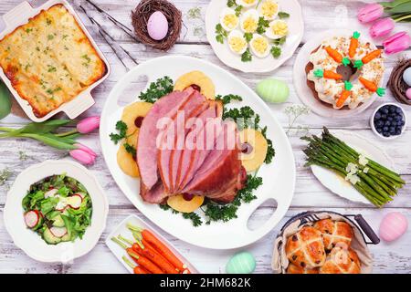 Traditional Easter ham dinner. Top view table scene on a white wood background. Ham, scalloped potatoes, eggs, hot cross buns, carrot cake and vegetab Stock Photo