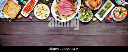 Traditional Easter ham dinner. Above view top on a dark wood banner background with copy space. Ham, scalloped potatoes, vegetables, eggs, hot cross b Stock Photo