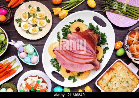 Traditional Easter ham dinner. Above view table scene on a dark wood background. Ham, scalloped potatoes, eggs, hot cross buns, carrot cake and vegeta Stock Photo