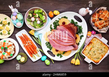Traditional Easter ham dinner. Top down view table scene on a dark wood background. Ham, scalloped potatoes, vegetables, eggs, hot cross buns and carr Stock Photo