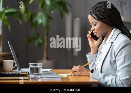 Serious afro woman in suit talking on mobile phone and writing down notes in cozy workplace Stock Photo