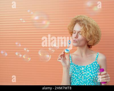 young cheerful curly redhead woman in blue sundress and straw hat with bubbles on beige background. Fun, summer, fashion, youth concept Stock Photo