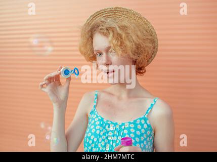 young cheerful curly redhead woman in blue sundress and straw hat with bubbles on beige background. Fun, summer, fashion, youth concept Stock Photo