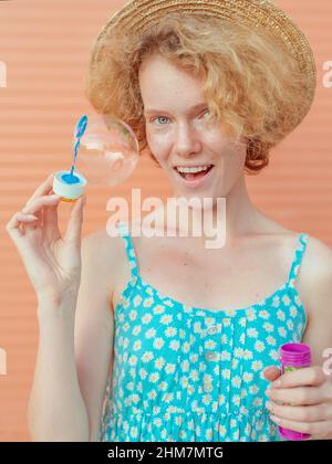 young cheerful curly redhead woman in blue sundress and straw hat with bubbles on beige background. Fun, summer, fashion, youth concept Stock Photo
