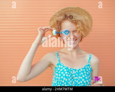 young cheerful curly redhead woman in blue sundress and straw hat with bubbles on beige background. Fun, summer, fashion, youth concept Stock Photo
