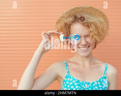 young cheerful curly redhead woman in blue sundress and straw hat with bubbles on beige background. Fun, summer, fashion, youth concept Stock Photo