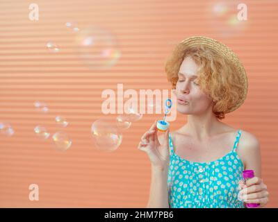 young cheerful curly redhead woman in blue sundress and straw hat with bubbles on beige background. Fun, summer, fashion, youth concept Stock Photo