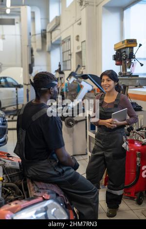 Vertical portrait of two smiling car mechanics chatting in garage shop, focus on young woman in workwear Stock Photo