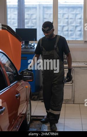 Vertical full length portrait of African-American mechanic standing by car in auto repair shop and inspecting vehicle Stock Photo