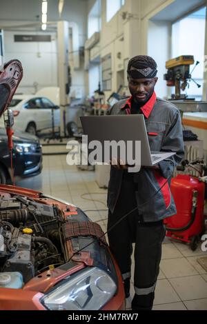 Vertical portrait of African-American mechanic standing by car in auto repair shop and using laptop while inspecting vehicle Stock Photo
