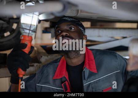 Portrait of African-American mechanic inspecting car on vehicle lift in auto repair shop, copy space Stock Photo
