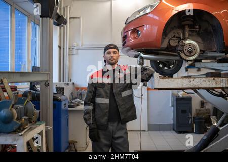 Front view portrait of Caucasian mechanic looking at camera while standing by car on lift in auto repair workshop, copy space Stock Photo