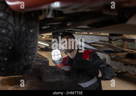Portrait of Caucasian mechanic inspecting car on vehicle lift in auto repair shop and holding portable worklight, copy space Stock Photo