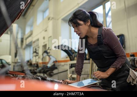 Side view portrait of young female mechanic looking under hood while inspecting vehicle in car repair shop, copy space Stock Photo