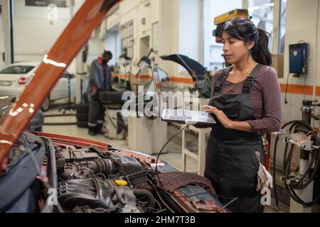 Side view portrait of young female mechanic using laptop while inspecting vehicle in car repair shop, copy space Stock Photo