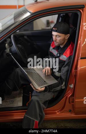 Vertical portrait of male mechanic using laptop while inspecting vehicle for engine errors in car repair shop Stock Photo