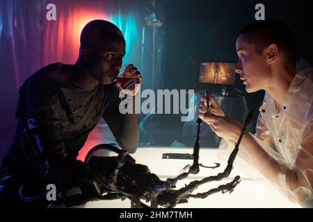 Woman making cyber arm and talking to African man sitting in front of her while he drinking alcohol Stock Photo