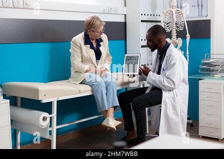 Specialist physician doctor holding tablet showing body radiography explaining bones pain to retired woman patient discussing healthcare treatment in hospital office. Anatomy skeleton on screen Stock Photo