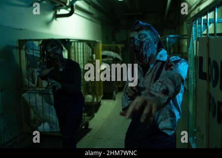 Portrait of gory zombie reaching out to camera while standing in dark industrial hallway, FX makeup, copy space Stock Photo
