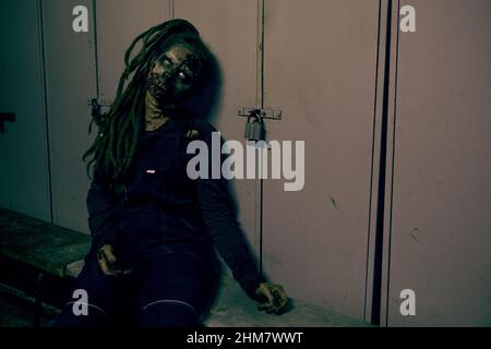 Shot of creepy female zombie sitting on floor by metal door lit by dark green light, copy space Stock Photo
