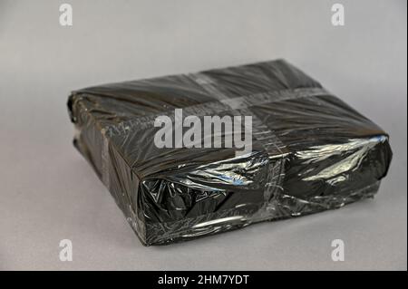 Black rectangular box wrapped in black film. The overlap is sealed with transparent adhesive tape.  Parcel packaging. Indoors. Selective Focus. Stock Photo