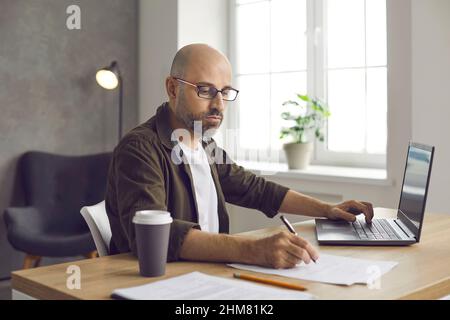 Serious man sitting at desk in home office, working on laptop computer and taking notes Stock Photo
