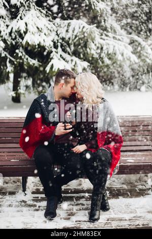 Couple in love sitting on bench and smiling in snowing forest. Stock Photo