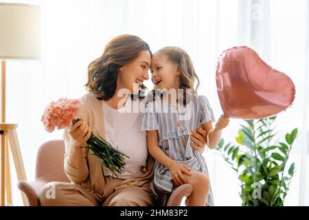 Happy day! Child daughter is congratulating mother and giving her flowers. Mum and girl smiling and hugging. Family holiday and togetherness. Stock Photo