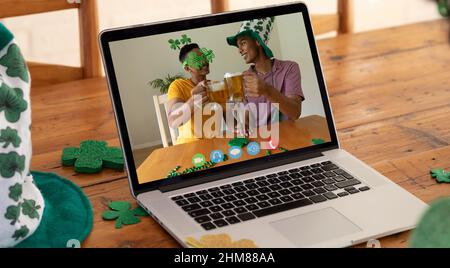 Webcam view of two african american men toasting beers on video call on laptop on wooden table Stock Photo