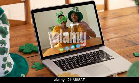 Webcam view of two african american men holding beers on video call on laptop on wooden table Stock Photo