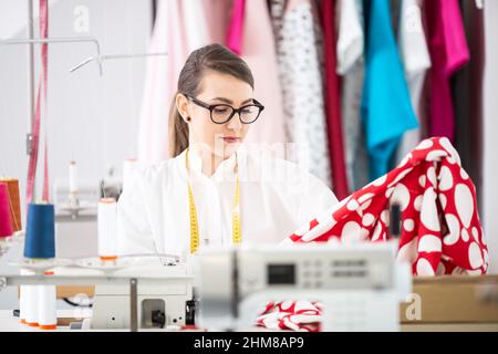 Tailor sweing dress in the workshop wearing classes and measuring tape around her neck. Stock Photo