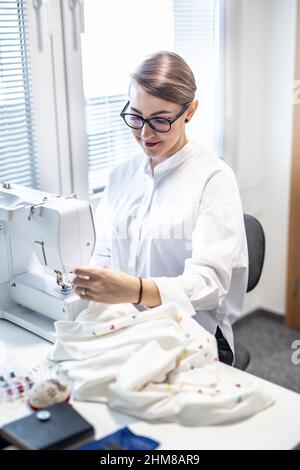 Smiling woman sews a blouse on a sewing machine at home, smiling. Stock Photo