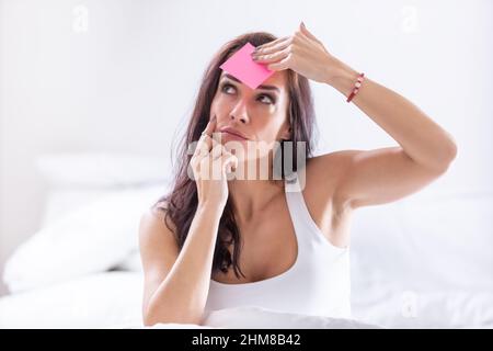 Woman forgetting something puts a pink post-it on her forehead, thinking. Stock Photo