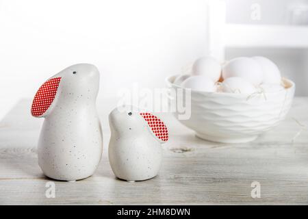 Two figurines of white hares with red ears big and small on a wooden saw cut and a bowl with eggs in the background in a white Scandinavian style kitc Stock Photo