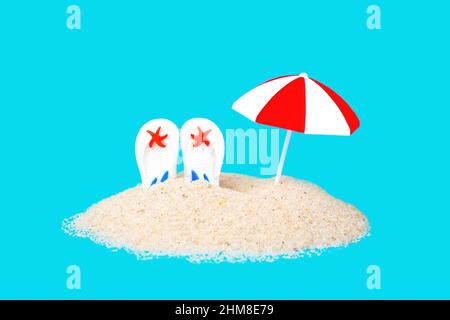Toy sun umbrella and white flip flops sticking out of a sand pile isolated on blue background imitating water. Creative beach vacations concept. Stock Photo