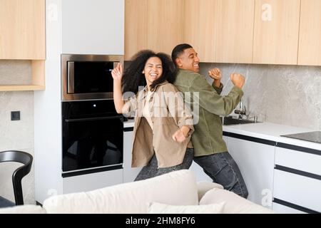 Waist up portrait view of the young couple in domestic clothes standing back to back to each other while dancing in the kitchen at home. Stock photo Stock Photo