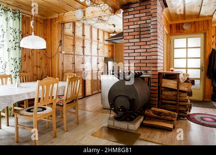 Bullerjan home wood burn stove heating device in wooden cottage home interior, with stack of firewood next to it. Kitchen table with chairs. Stock Photo