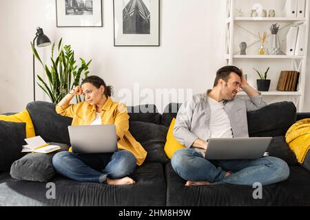 Tired from work on the distance middle-age couple sitting separate on the couch with laptops and looking in different sides, unemployed spouses has financial problems, wife and husband don't talk Stock Photo