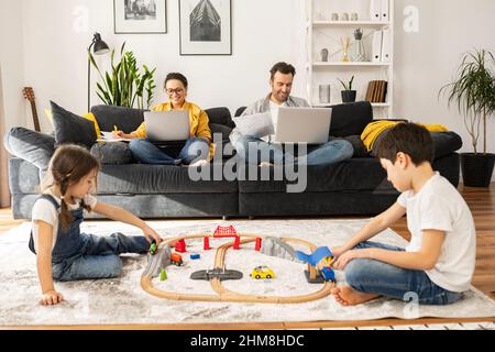 Busy parents work remotely with laptops sitting on the couch in living room, two sweet kids playing with toys on the foreground, freelancer parents working on the distance during quarantine Stock Photo