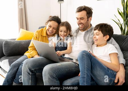 Cheerful happy family of four using laptop sitting on the sofa in cozy apartment, parents and two kids watching movie on computer, comedy, spends time on the weekend together at home Stock Photo