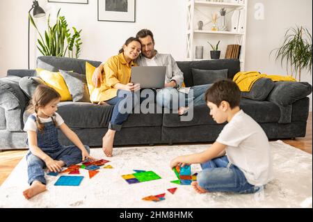 Happy family of four spends weekend at home. Parents using laptop sitting on the sofa in cozy living room, watching movies while two kids playing with constructor on the floor. Family idilia Stock Photo