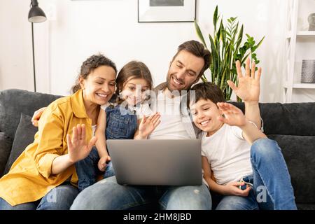 Family of four using laptop for video connection with grandparents or family. Multiracial mom, dad and two kids waving in webcam of the laptop, video call, virtual meeting concept Stock Photo