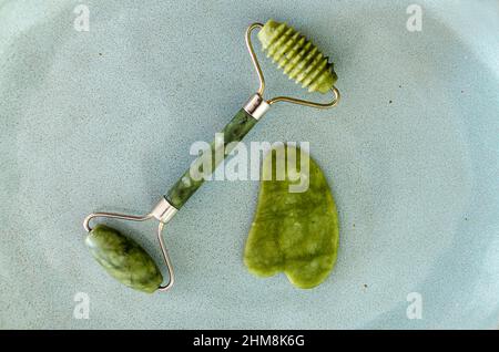 Green face roller and gua sha massager. Jade roller isolated on blue background. Facial massage concept. Stock Photo